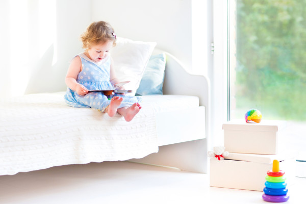 Adorable toddler girl reading a book sitting on a white bed at a